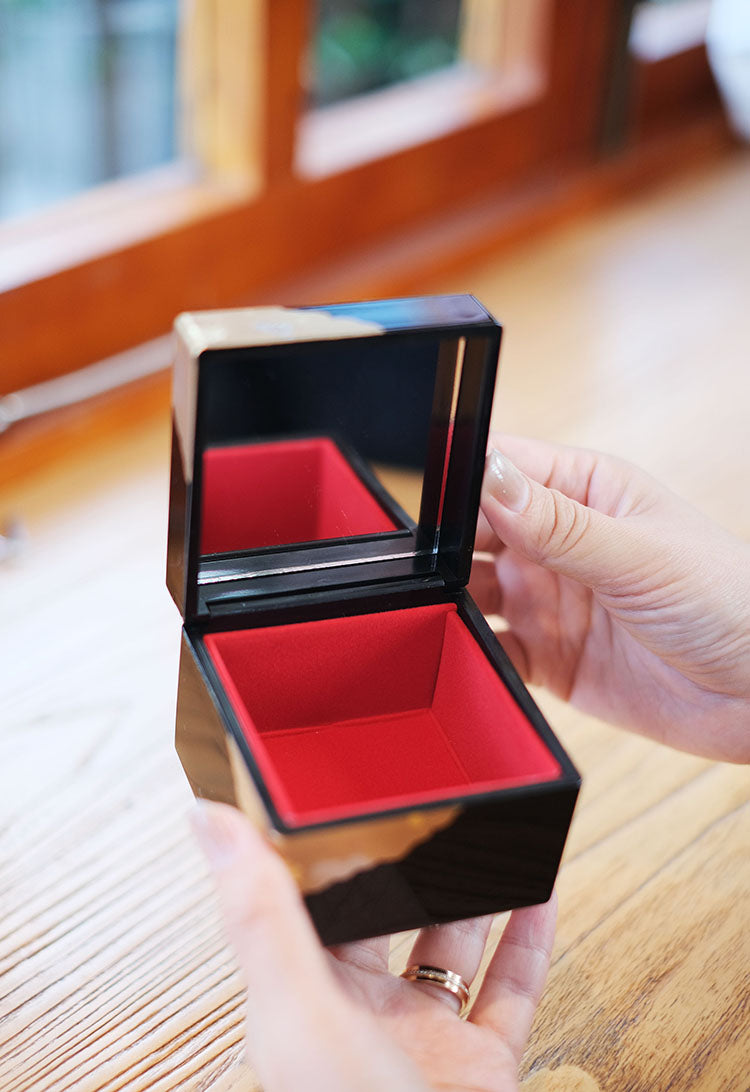 Japanese lacquerware jewelry box open showing black glossy exterior, red velvet interior and mirror held in hands on wooden table