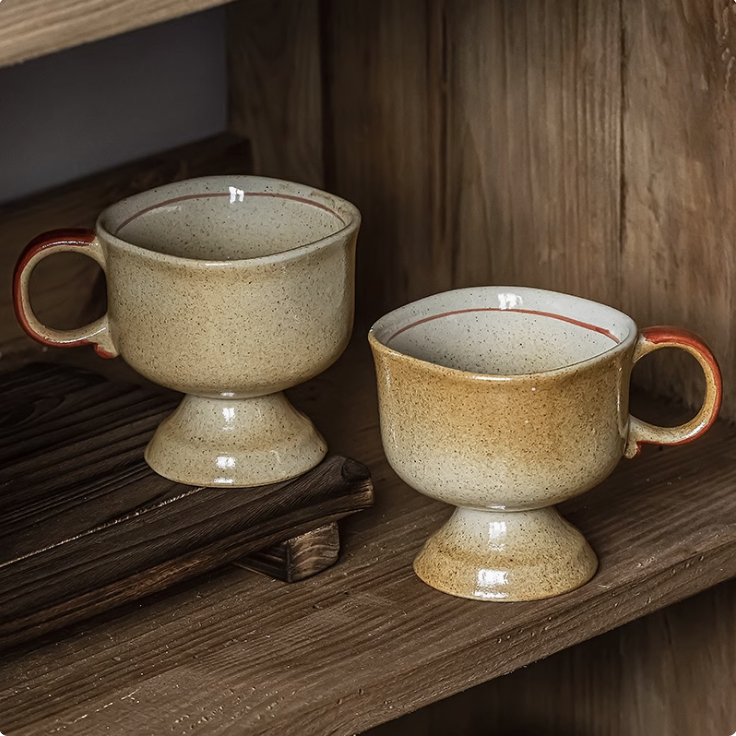 Two beige speckled Japanese vintage ceramic cup pedestal mugs with red rim and handle on wooden shelf