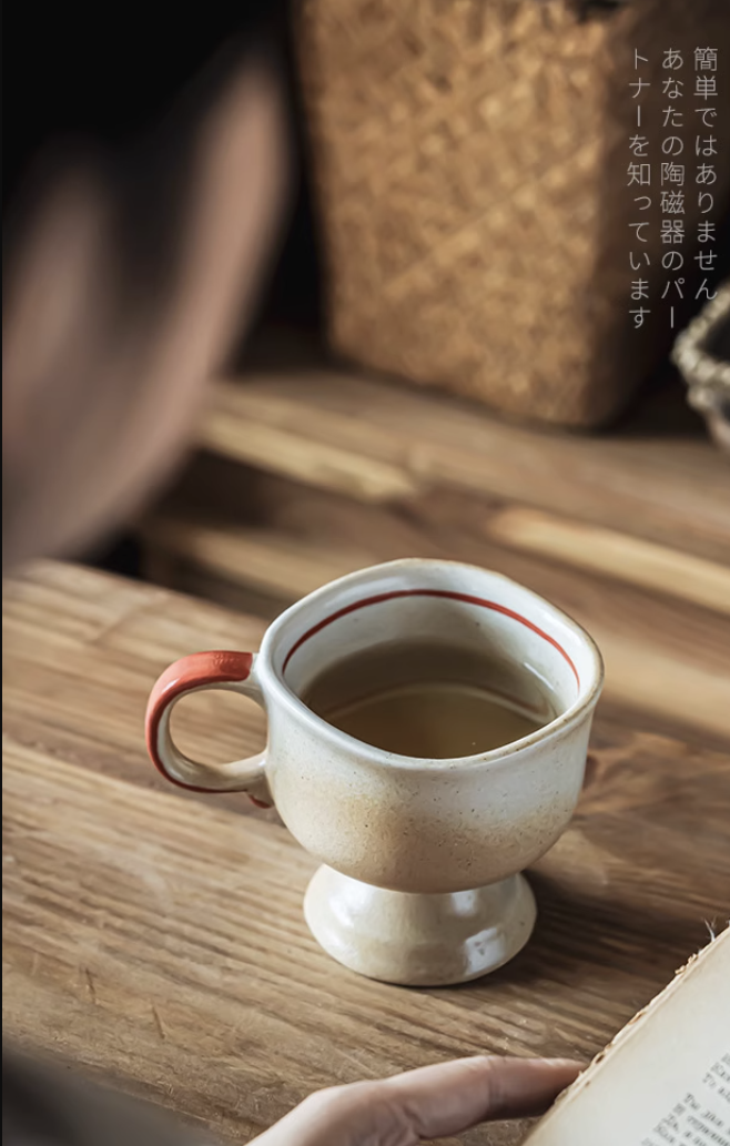 Japanese vintage ceramic cup with red rim and handle filled with tea on wooden table next to open book