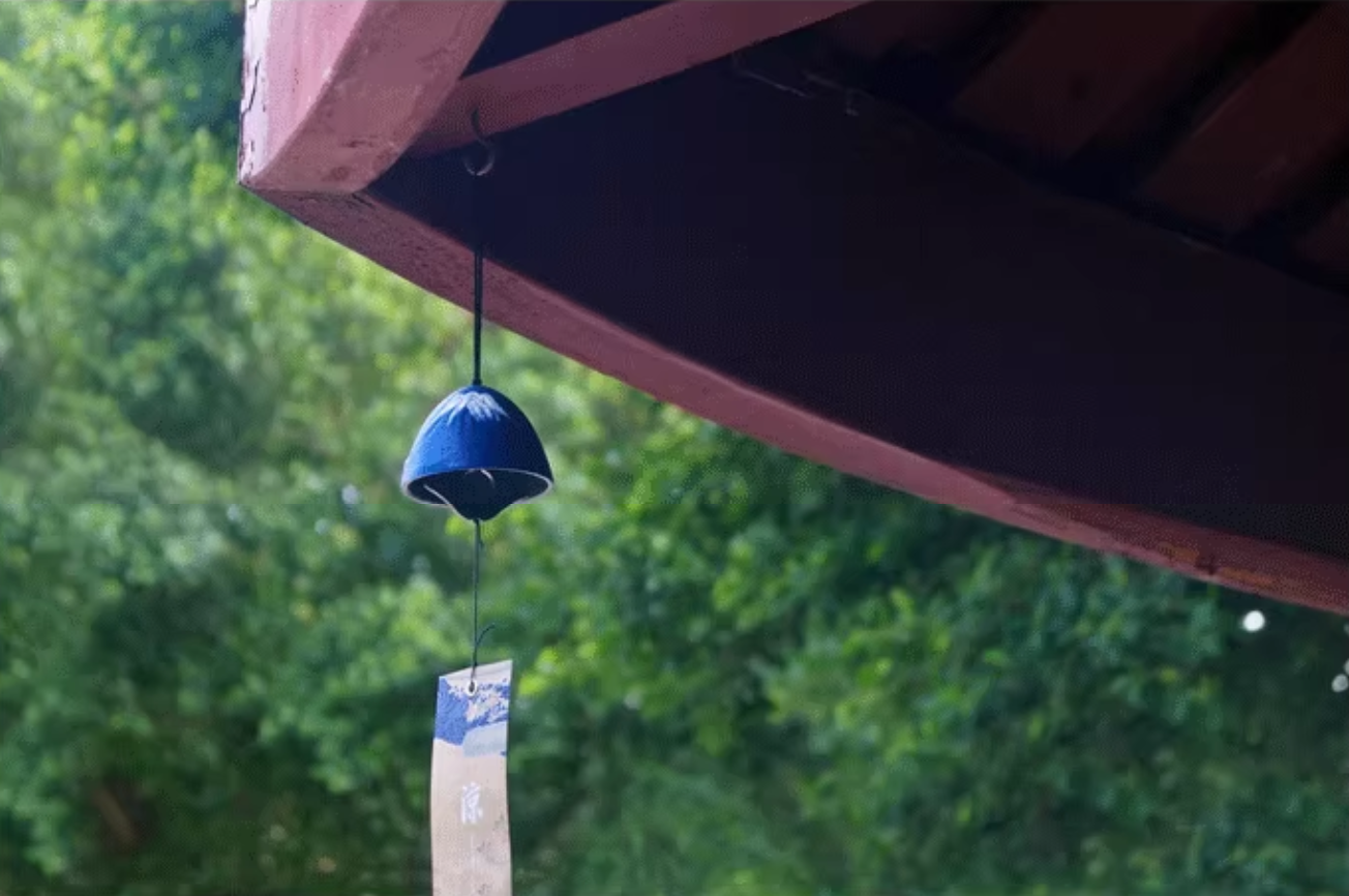 Rin Essense blue Japanese ceramic wind chime hanging from porch eave with paper strip and blurred trees