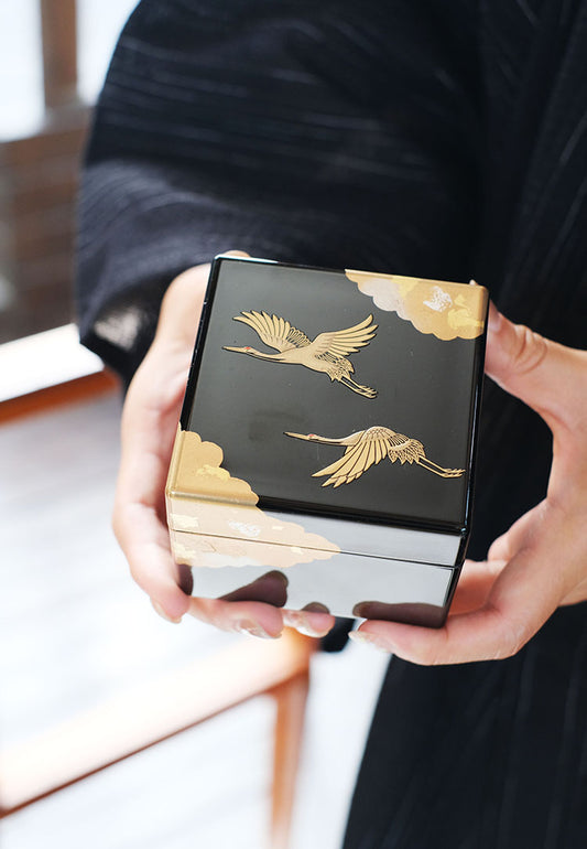 Hands holding Rin Essense black Japanese lacquerware jewelry box with gold crane and cloud makie design