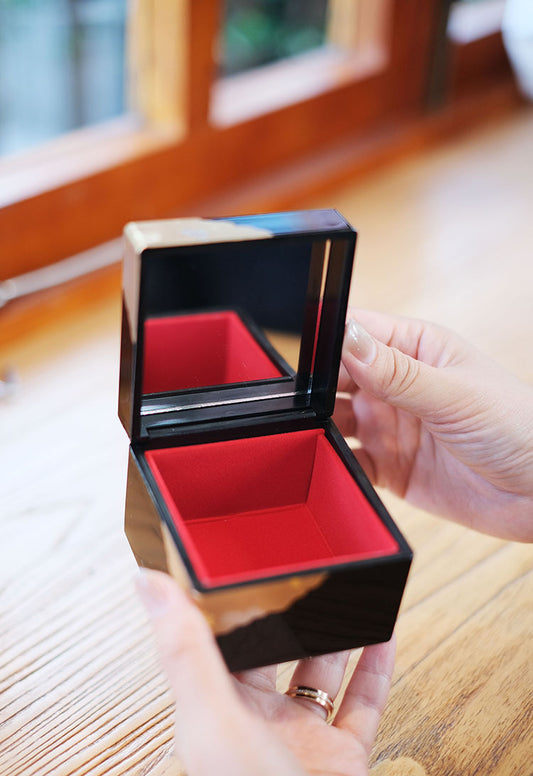 Japanese lacquerware jewelry box open showing black glossy exterior, red velvet interior and mirror held in hands on wooden table
