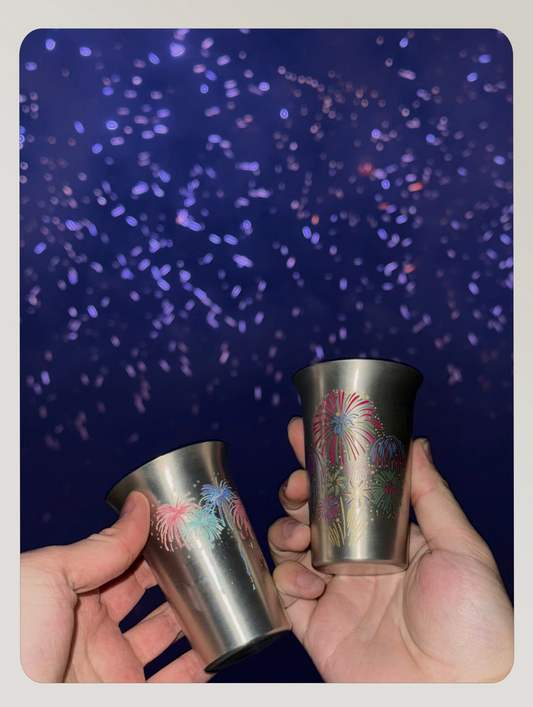 Two hands holding Rin Essense stainless steel sake cup sets with colorful fireworks design against a dark blue background