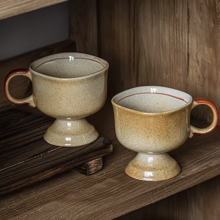 Two beige speckled Japanese vintage ceramic cup pedestal mugs with red rim and handle on wooden shelf